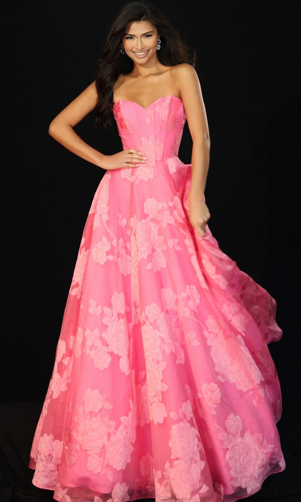 A woman smiles against a black background, wearing the Jovani 47327 long pink prom dress with a strapless corset bodice and subtle floral pattern.